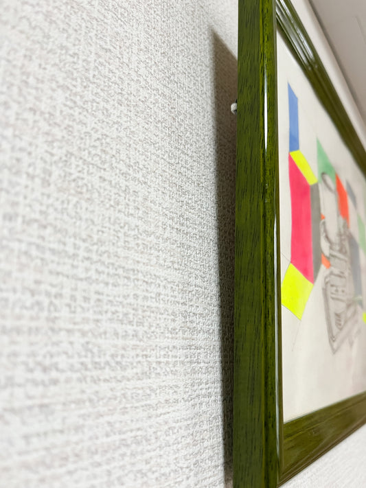 光藤有佳のデッサン画を斜めから撮影。モスグリーンの額縁と、ケーキセット＋ジオメトリック背景が立体的に見える。Angled view of Yuka Mitsufuji’s framed drawing. Moss green frame, cake still life, and vivid geometric background in perspective.