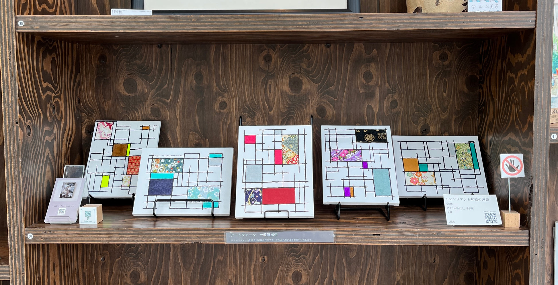 兵庫県神戸市の図書館兼コンサートホールの壁面ギャラリーに展示された、光藤有佳（Yuka Mitsufuji）による幾何学模様と和紙のコラージュ作品。5点が並んだ展示風景で、壁にかけてもスタンドに置いても飾れるアート。Exhibition view at the library and concert hall in Kobe, Japan, featuring five geometric collage artworks with washi paper by Yuka Mitsufuji. Displayed on a wall gallery, the pieces can be showcased both hanging and standing, ideal for modern interior decor.